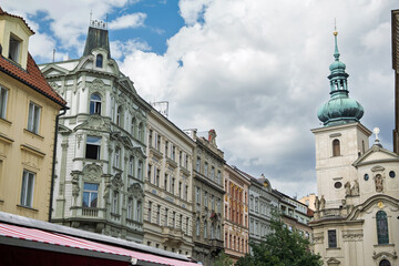 Obraz premium A View Of Tall, Colorful Historic Buildings In Prague With An Ornate Church Tower And Spire Reaching Towards A Blue Sky With White Clouds, Czech Republic