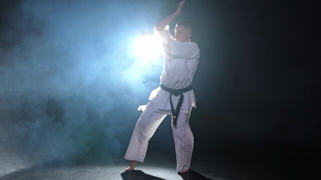 Strong man fighter in white kimono for karate martial arts with black belt preparing for fight training in black background. Man training moves punches kicks practicing shadow fight ready for sparring