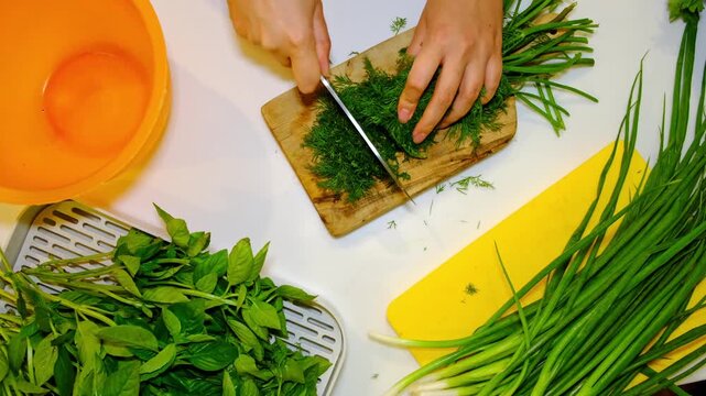Top view of hands finely chopping parsley on a wooden board, surrounded by basil, dill and green onions on a bright kitchen table. Fresh herbs support healthy home cooking and mindful eating.