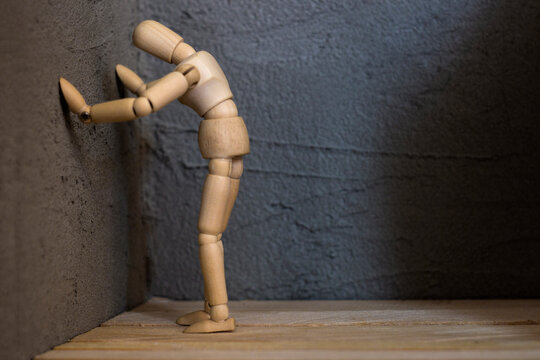 A wooden mannequin rests its hands against a gray concrete wall. Its hunched posture conveys heaviness, impotence, or deep remorse. Dramatic minimalism.