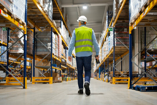 A worker in a safety vest and hard hat walks down the aisles of a warehouse. Tall shelves filled with boxes line both sides. The setting is bright with overhead lights.