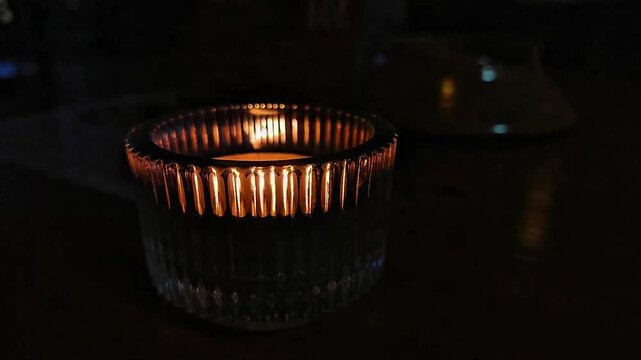 A lit candle in a glass container on a table in an outdoor restaurant sways in the night wind.