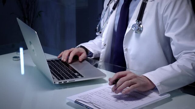Medical doctor in lab coat with stethoscope typing on laptop in a dim office, managing electronic health records and telemedicine data for modern digital healthcare workflows