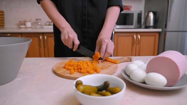 The chef prepares Olivier salad. Caucasian chef carefully slices carrots. Kitchen cook chopping vegetables next to eggs, pickles, and boiled sausage on a cutting board in the kitchen