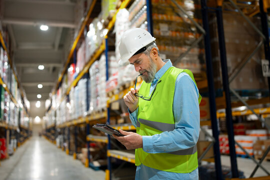 A worker wearing a hard hat and safety gear stands in a warehouse. He is holding a clipboard and looking at the items on the shelves. It is likely a busy time in the warehouse.