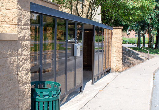 Modern glass bus stop shelter with sturdy stone wall, vibrant green trash can, and sloped steps with railing on a bright sunny day, reflecting the bustling street, a car, and lush green trees.
