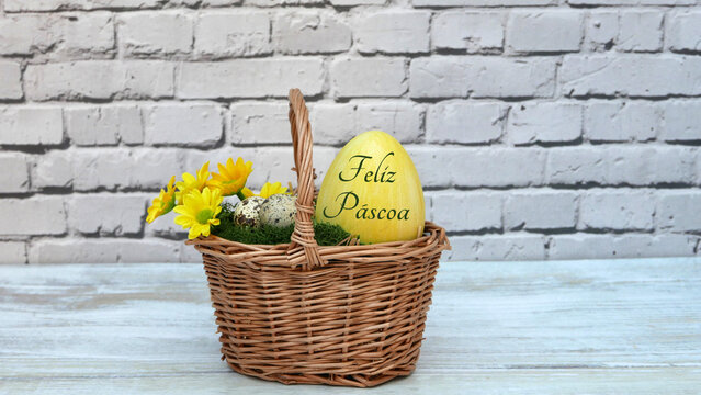 A sauda&ccedil;&atilde;o portuguesa de P&aacute;scoa "Feliz P&aacute;scoa" escrita num ovo amarelo dentro de um cesto de vime em frente a uma parede de tijolos brancos.