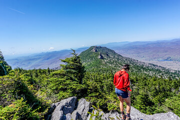 Hiker stands atop rocky peak, gazing at distant mountain range © ExploringandLiving
