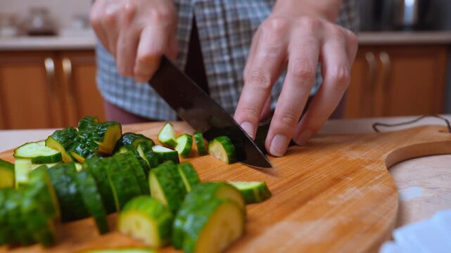 Female hands cutting cucumber on wooden board, sharp knife slicing fresh green cucumber, close-up of smooth movement, plaid shirt sleeve, kitchen counter, healthy salad preparation, morning