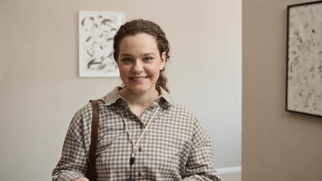 Tilt up slowmo portrait of young Caucasian woman taking off audio guide earphones and smiling at camera, posing in modern art gallery with framed abstract paintings in blurred background