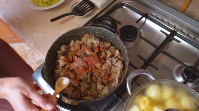 Cooked meat with tomatoes and spices mixed in a skillet, hands spoonful of paprika and seasoning into hot meat, bubble sauce and steam, cozy stovetop cooking for a hearty dinner