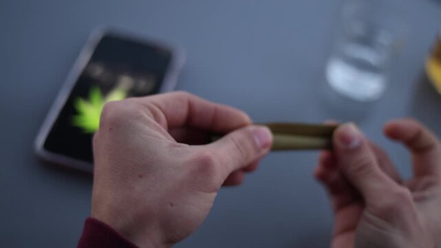 Close up of hands rolling a marijuana blunt with a cannabis leaf symbol on a smartphone nearby