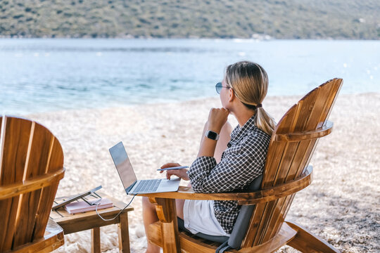 Young woman working on laptop by the sea with portable solar charger, digital nomad and workation lifestyle, remote work, sustainable travel and beach office concept.