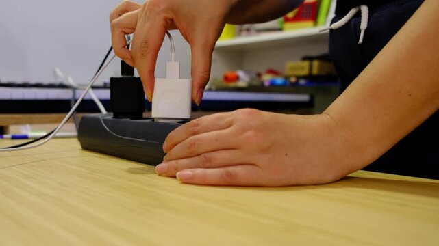 Close shot shows hands unplugging each charger one by one from a power strip, cables looping over a wooden desk in soft indoor light. Cutting standby power saves energy and lowers fire risk.