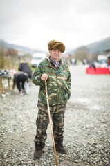 Naklejka premium A wise elder in camouflage gear and a furry hat stands confidently with a cane. Busy vendors and shoppers fill the vibrant market around him, set against picturesque hills