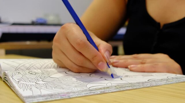 Close up shows hands coloring a detailed drawing with a blue pencil on a desk, shallow focus and calm indoor light. Adult coloring reduces stress, supports mindfulness, and helps prevent burnout.