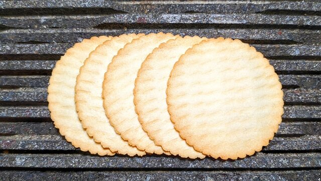 Top view of five thin, round potato biscuits with scalloped edges, arranged in a row on a dark, textured stone background. Crispy golden savory crackers for a snack or appetizer.