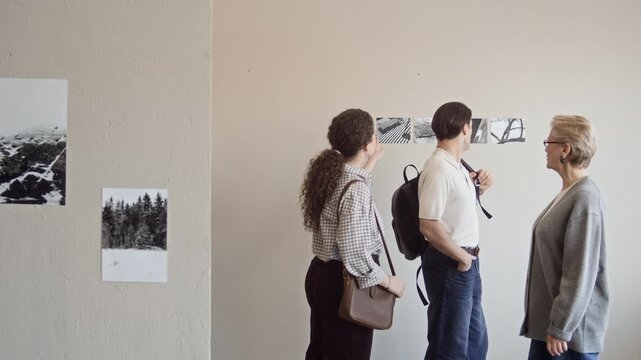 Tracking shot of confident mature Caucasian woman guiding young couple of gallery visitors through exhibition of black and white fine art photography