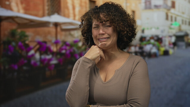 Hispanic woman covering face with hand at outdoor restaurant terrace beside flower pots and tables; sadness.