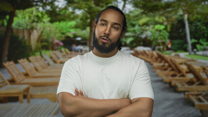 Man with beard and arms crossed wearing white tshirt by poolside lounge chairs among tropical trees...