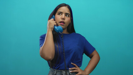 Young hispanic woman holding blue telephone handset to ear with hand on hip in studio, wearing blue t shirt and jeans  pensive. © Krakenimages.com
