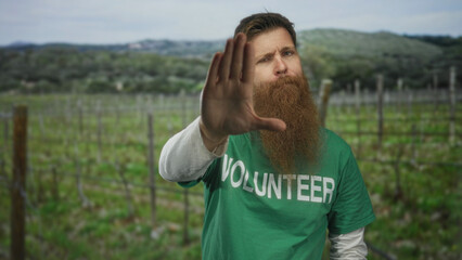 Naklejka premium Man wearing green volunteer shirt raises hand in stop gesture in a countryside field; safety awareness.
