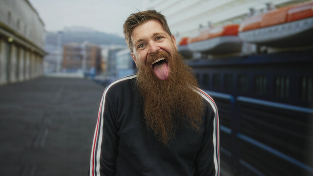 Bearded man leans on railing by lifeboat and winks eye and sticks tongue out on cruise ship deck; lighthearted fun.