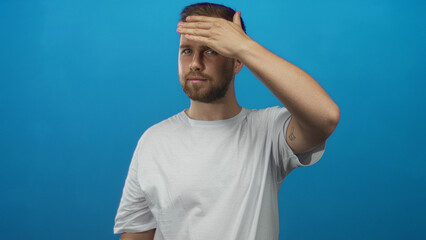 Naklejka premium Man with beard and white t shirt covering his eyes with one hand in a blue studio, face partly hidden and anonymous; shyness.