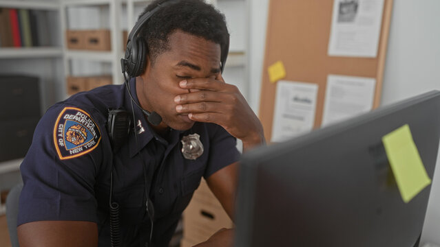 Man police officer in uniform wearing headset covers face with hand at desk by computer monitor and corkboard in building; stress.