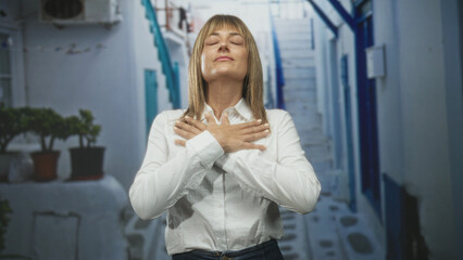 Fototapeta premium Woman in white shirt with hands crossed on chest, eyes closed, head tilted back, standing before whitewashed steps and potted plants in a narrow street; serenity mindfulness.
