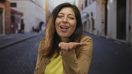 Fototapeta premium Woman in tan jacket blows kiss with hand to lips in old town street smiling warmly; affectionate greeting.
