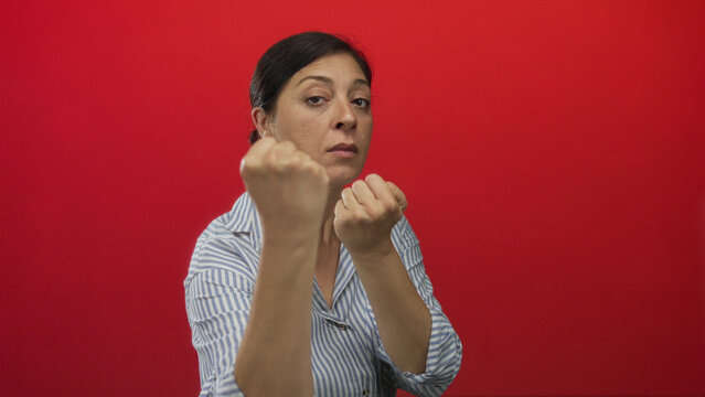 Middle aged woman in striped shirt with bare fists raised in fighting stance in red studio setting; defiance.