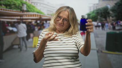 Fototapeta premium Woman holds blue medicine bottle in outstretched hand at busy outdoor market stall, smiling and presenting pills; wellness comfort.