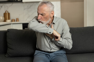 Fototapeta premium Elderly man coughing into elbow feeling unwell on sofa at home