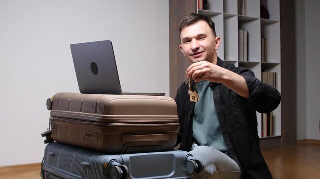 Man holding a tiny house keychain with keys while sitting next to stacked luggage and a laptop, symbolizing relocation, new beginnings, and searching for real estate