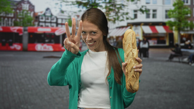 Young hispanic woman holding a baguette and showing three fingers on a city street while smiling, wearing green cardigan and white top; joy community.