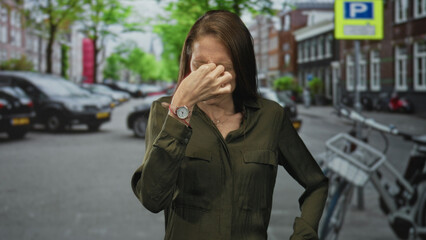 Fototapeta premium Young hispanic woman pinches nose with watch visible on a city street near parked cars and a bicycle rack and trees by buildings; disgust.