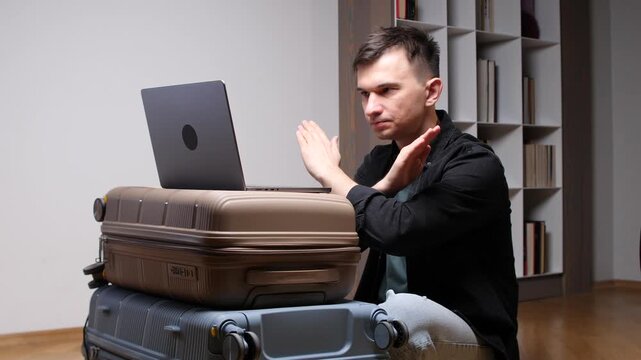 Man crossing hands in a stop gesture while looking at a laptop computer balanced on stacked suitcases, indicating rejection of travel plans or a canceled trip
