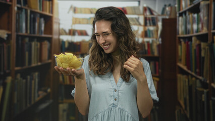 Woman covers mouth with hand while holding a transparent bowl of potato chips among tall bookshelves in library  fun moment. © Krakenimages.com