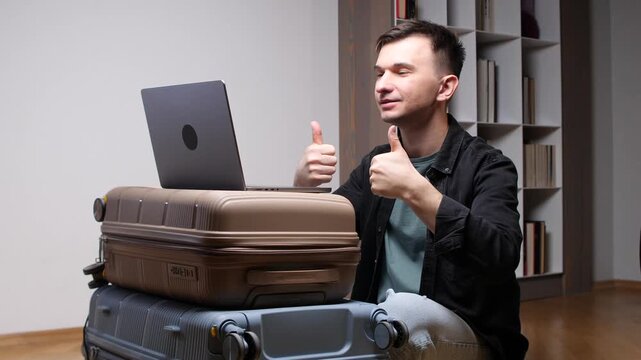 Young man with luggage gives two thumbs up while using a laptop indoors, celebrating successful online booking and vacation planning before departure, smiling and satisfied
