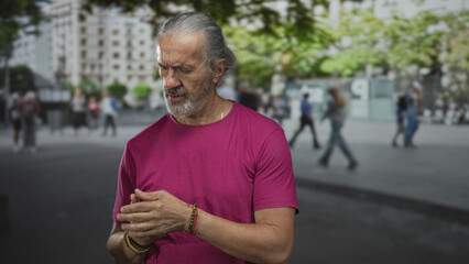 Fototapeta premium Man with clasped hands and bracelets, gray hair pulled back, looking down on a busy urban street; thoughtful reflection.