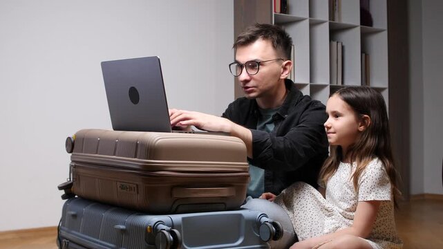 Father booking travel and looking at screen with his daughter, sitting next to a stack of suitcases, preparing for a family trip using modern technology at home