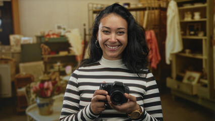 Woman holding camera and looking to the side in a home decor shop interior, striped sweater and hoop earrings visible, hands on camera  confidence. © Krakenimages.com