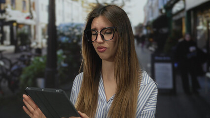 Woman wearing glasses holding tablet frowning on busy city street among blurred storefronts  frustration. © Krakenimages.com