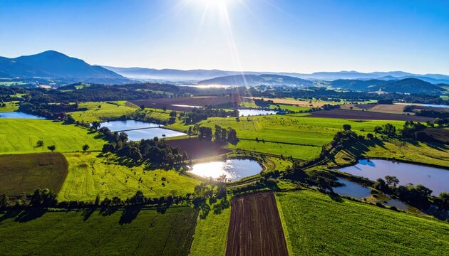 Expansive aerial view of a diverse agricultural landscape with green fields, planted crops, sparkling water bodies, and distant mountains under a radiant morning sun.