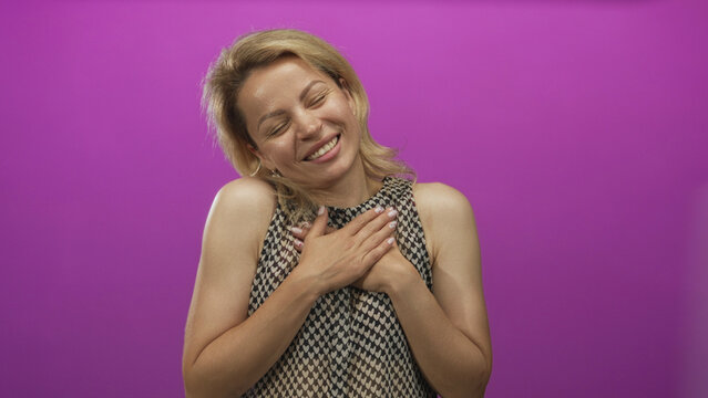 Young blonde woman touching chest and smiling warmly at camera in purple studio; gratitude kindness sincerity.