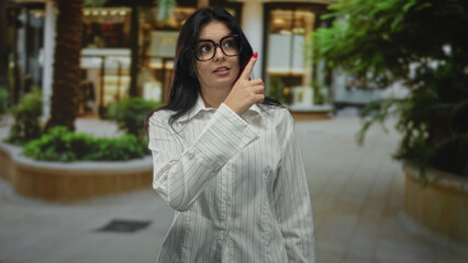Obraz premium Woman wearing glasses points finger to the right on urban street lined with shops; curiosity insight.