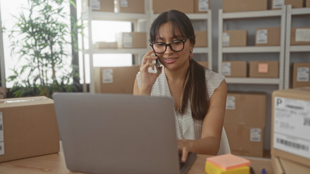 Hispanic woman typing on laptop and talking on phone amid parcel boxes in a building; small business focused.