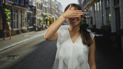 Obraz premium Young latina brunette woman hand covering eyes while smiling on a city street with glasses visible; playful curiosity.