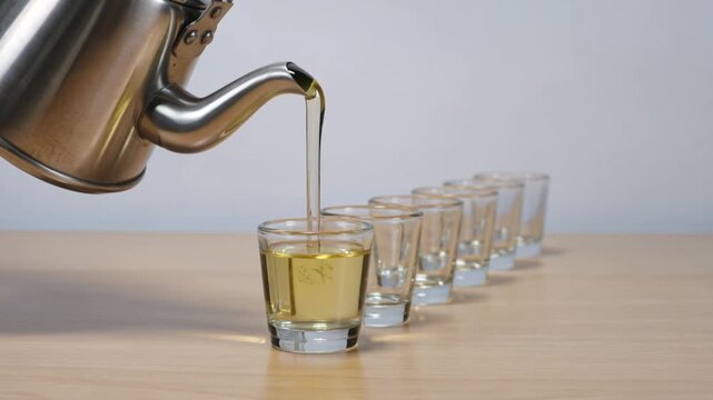 Pouring Tea from a Metal Kettle into Row of Glass Cups